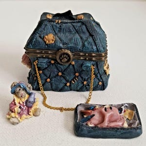 May include: A dark blue quilted handbag figurine with gold-tone hardware, a paw print clasp, and a gold chain strap. Beside it sits a small mouse figurine wearing a blue hat and pink dress, and a separate open dark blue rectangular case.