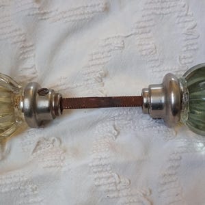 May include: Antique glass door knobs with a faceted design. The knobs are a pale yellow-green color, with silver-toned metal hardware and a rusty connecting rod. A vintage home decor item.