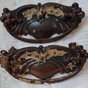May include: Two antique drawer pulls with a dark bronze finish and ornate detailing. The pulls have a curved design with a central decorative element and a ring handle. The paint is chipped, revealing the metal underneath, giving them a vintage look.