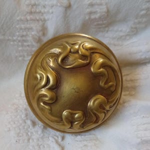 May include: A round, antique brass-colored knob with an ornate, swirling floral design. The knob has a slightly domed shape and a smooth, polished finish. The decorative element is raised and detailed.