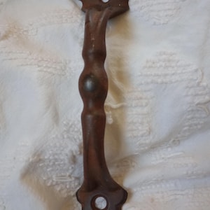 May include: A vintage, ornate, cast iron door handle with a keyhole. The handle is a dark, rusty brown color and features decorative floral-like details at both ends. The handle is approximately 8 inches long.