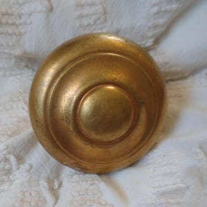 May include: A round, antique brass door knob with concentric circles. The knob has a warm, golden hue and a slightly aged appearance, suggesting it is vintage or has been well-used. The design is simple yet elegant.