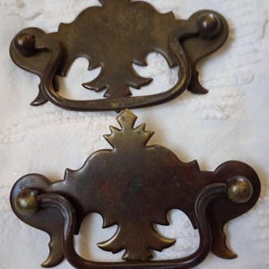 May include: Two antique bronze drawer pulls with ornate, stylized designs. Each pull features a decorative backplate with a central flourish and curved handle. The pulls have a dark, aged patina, suggesting vintage craftsmanship.