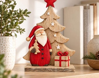 May include: A wooden tabletop decoration featuring a Santa Claus figure in a red coat and hat standing next to a natural-toned wooden Christmas tree. The tree is topped with a red star and small white stars. A gift box with a red bow sits at the base.