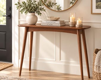 May include: A mid-century modern wooden console table with tapered legs and a curved apron. On the surface sits a textured beige vase with green foliage, a shallow bowl, two stacked books titled The Kinfolk Home, and two glass candle holders.