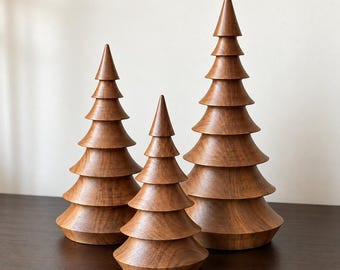 May include: Three conical wooden sculptures shaped like evergreen trees, each featuring a smooth, polished brown finish with visible wood grain patterns and tiered, horizontal layers that taper to a pointed top in varying heights.