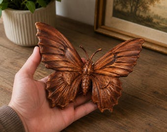 May include: A carved wooden butterfly sculpture featuring a rich, dark brown wood grain finish with detailed wing patterns, a central body, and two antennae, showcasing natural textures and a symmetrical shape in a medium-sized decorative form.