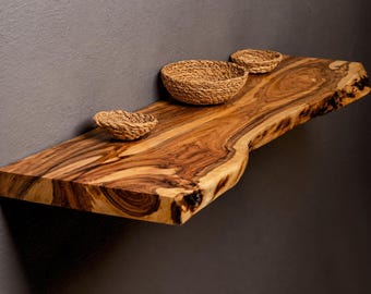 May include: A live edge wooden floating shelf with a prominent brown wood grain pattern and natural bark edges. Three small, round, woven seagrass bowls are placed on the surface of the shelf, varying in size from small to medium.