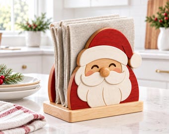 May include: A wooden napkin holder featuring a Santa Claus face design with a red hat, white beard, and natural wood base. The holder contains several folded beige linen napkins, standing upright on a flat, light-colored wooden rectangular base.