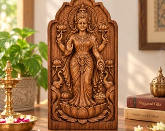 May include: A brown wooden wall plaque featuring a detailed relief carving of the Hindu goddess Lakshmi standing on a lotus flower, flanked by two elephants, with four arms, ornate jewelry, and coins flowing from one hand into a pot below.