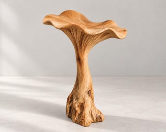 May include: A light brown wooden sculpture shaped like a mushroom with a wide, wavy cap and a thick, textured stem. The wood grain is visible throughout the piece, showing natural patterns and knots in the material, standing upright on a flat base.
