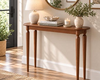 May include: A narrow wooden console table with four tapered legs, topped with a textured table lamp, a scalloped bowl, two stacked books titled The Kinfolk Home and At Home, a glass candle, and a ceramic vase filled with green leafy branches.