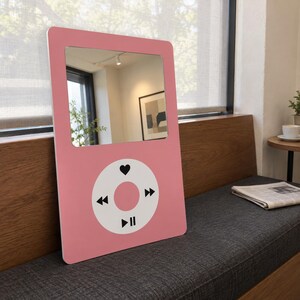 May include: A rectangular pink mirror designed to resemble a vintage digital music player. The top section features a reflective glass surface, while the bottom section displays a white circular control interface with black icons for play, pause, skip, and a heart symbol.