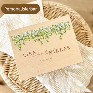 May include: A rectangular light-colored wooden box featuring a printed illustration of hanging white flowers and green leaves at the top. The text reads LISA und NIKLAS with the date 12.06.2027 printed below in a brown sans-serif font.