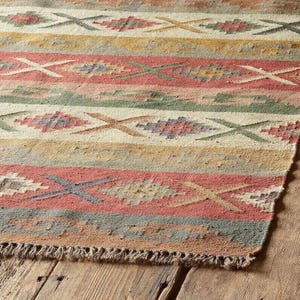 Può includere: Un tappeto intrecciato con fasce orizzontali nei colori rosso spento, crema, giallo e verde salvia. Il motivo è composto da forme geometriche a rombo e motivi a X ripetuti sulla superficie, rifinito con una corta frangia sul bordo inferiore.