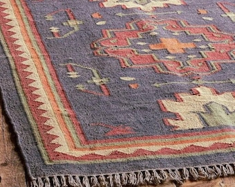 May include: A rectangular area rug featuring a deep purple base with geometric patterns in cream, sage green, and burnt orange. The design includes a central stepped diamond motif and a zigzag border, finished with a short, knotted fringe edge.