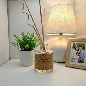 May include: A clear glass reed diffuser bottle with a label reading Home is where the Heart is, containing wooden reeds. Next to it are a small green artificial plant in a white pot, a white table lamp with a textured shade, and a light wood photo frame.
