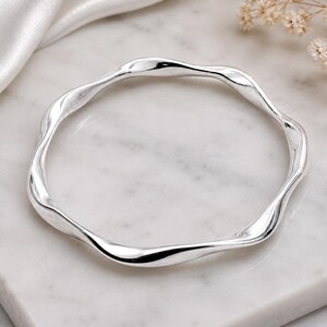 May include: A polished silver bangle bracelet featuring a continuous wavy, undulating design. The metal surface is smooth and reflective, creating a series of gentle curves that form a circular shape. The bangle has a high-shine finish.