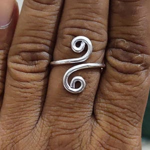 May include: A silver-colored metal ring featuring a bypass design with two symmetrical spiral ends. The band is constructed from a single piece of wire that curves into two distinct circular coils facing opposite directions on the finger.