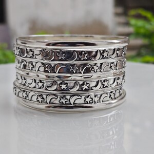 Peut inclure: Une pile de bracelets rigides en métal argenté présentant un motif en relief répétitif de croissants de lune et d'étoiles à cinq branches. Les bracelets ont une finition polie avec des zones en creux assombries pour souligner les motifs.