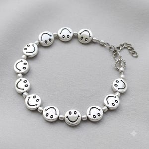 Könnte beinhalten: Ein silberfarbenes Armband mit einer Reihe runder, flacher Perlen, die jeweils mit einem schwarzen Smiley-Gesicht geprägt sind. Das Armband enthält kleine kugelförmige Zwischenperlen und eine Metallkettenverlängerung mit einem Karabinerverschluss.