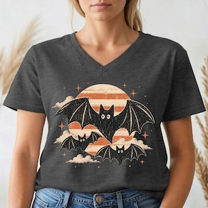 May include: A dark heather gray V-neck t-shirt featuring a graphic print of three black bats flying in front of a large, orange, striped moon. The design includes small orange stars and white clouds surrounding the bats, creating a nocturnal scene.