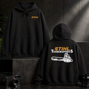 Op de afbeelding: Een zwarte hoodie met het oranje STIHL-logo op de borst. De achterkant toont het oranje STIHL-logo boven de witte tekst TIMBERSPORTS en een gedetailleerde witte grafische illustratie van een kettingzaag met een lang zaagblad.
