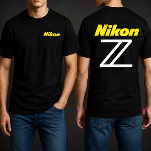 Puede incluir: Una camiseta negra de manga corta con el logotipo amarillo de Nikon en el pecho izquierdo. La parte posterior de la camiseta muestra el logotipo amarillo de Nikon sobre un gráfico grande de la letra Z en blanco. Se combina con vaqueros azules.