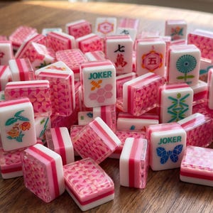 May include: A collection of pink and white Mahjong tiles featuring various designs including the word JOKER with a butterfly or honeycomb graphic, floral patterns, Chinese characters, and geometric textures on the backs of the rectangular pieces.