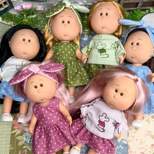 Könnte beinhalten: Eine Gruppe von sechs Puppen mit verschiedenen Haarfarben und Stilen, die Outfits wie gepunktete Kleider, Oberteile mit Mausmotiven und passende Stirnbänder tragen. Jede Puppe hat ein rundes Gesicht, kleine Gesichtszüge und weiße Schuhe.