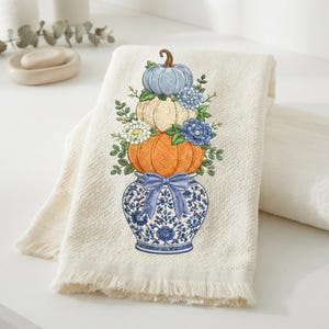 May include: A cream-colored hand towel with a fringed edge featuring an embroidered design of a blue and white patterned vase holding a stack of three pumpkins in blue, cream, and orange, accented with blue and white flowers and green leaves.