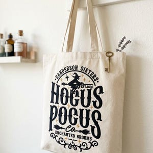 May include: A cream-colored canvas tote bag featuring a black and gold graphic print that reads Sanderson Sisters Hocus Pocus Co. Est. 1693 Enchanted Brooms, with a silhouette of a witch on a broomstick and a small metal key attached to the strap.