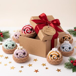 Puede incluir: Una caja de regalo de cartón marrón con una cinta de satén roja y una etiqueta que dice Merry Christmas, rodeada de varios pequeños dulces de ganchillo en cestas marrones, cada uno con una parte superior en espiral, ojos negros y sonrisa.