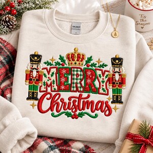May include: A beige crewneck sweatshirt featuring an embroidered design with the text Merry Christmas in red and green. The graphic includes two nutcracker figures, a gold crown, green holly leaves, and small gold stars on the front.