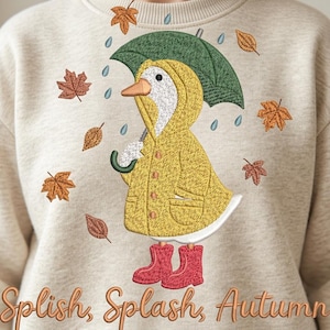 May include: A beige crewneck sweatshirt featuring an embroidered design of a white duck wearing a yellow raincoat and red rain boots, holding a green umbrella. Falling autumn leaves and raindrops surround the duck, with the text Splish, Splash, Autumn.