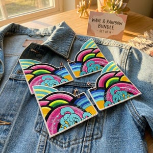 May include: Four embroidered patches featuring a wave and rainbow design in vibrant pink, yellow, blue, and green threads. Each patch is shaped like a quarter-circle with a white border and a small metal safety pin attached to the fabric surface.