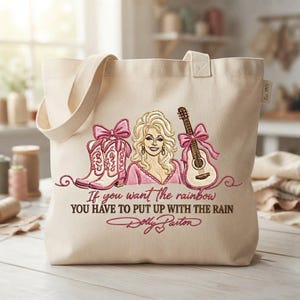 May include: A beige canvas tote bag featuring an embroidered design of Dolly Parton, pink cowboy boots with bows, and an acoustic guitar. The text reads: If you want the rainbow YOU HAVE TO PUT UP WITH THE RAIN, followed by the signature Dolly Parton.