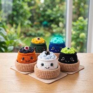 May include: Five small, round, crocheted plush figures with smiling faces, each sitting in a tan crocheted bowl. The figures are orange, dark green, blue, white, and black, topped with textured yarn and a single plastic eye in various colors.