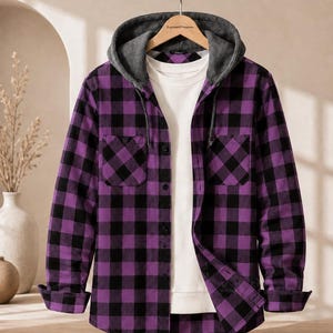 Può includere: Una camicia a maniche lunghe con bottoni, caratterizzata da un motivo a quadri viola e nero, un cappuccio grigio con coulisse e due tasche sul petto. La camicia è sovrapposta a una t-shirt bianca semplice ed è appesa a una gruccia in legno.
