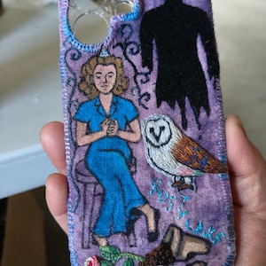 May include: A purple fabric phone case featuring hand-embroidered designs including a person in a blue outfit, a black silhouette figure, a barn owl, a fallen pot with a rose, and the text Rusty Lake stitched in light blue thread.