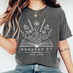 May include: A charcoal gray short-sleeve t-shirt featuring a white line art graphic of an open book with wildflowers growing from the pages. The text reads chapter 57 and EST. 1969, centered below the book illustration in a clean serif font.