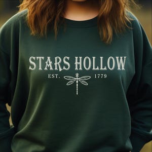 May include: A dark forest green crewneck sweatshirt featuring the white text STARS HOLLOW centered on the chest. Below the text, the word EST. appears to the left and 1779 to the right of a white line-art illustration of a dragonfly.