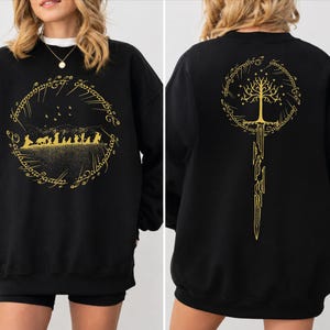 May include: A black long-sleeve sweatshirt featuring gold graphic prints. The front displays a silhouette of a group of figures within a circular Elvish script border. The back features a gold tree and a broken sword inside a circular Elvish script.