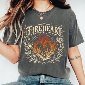 May include: A charcoal gray short-sleeve t-shirt featuring a graphic print of a flaming heart surrounded by floral vines and stars. The text FIREHEART is printed in a decorative cream-colored font above the heart, which is colored in orange and red.