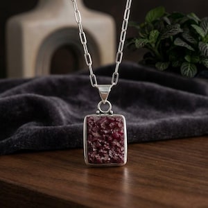 May include: A silver necklace featuring a rectangular pendant with a deep red, textured druzy stone center. The pendant hangs from a silver chain composed of elongated, oval-shaped links, creating a clean and structured appearance for the jewelry piece.