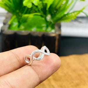 Peut inclure: Une bague de couleur argentée présentant un motif répétitif et continu de symboles infinis qui entoure tout l'anneau. Le métal possède une finition lisse et polie avec des espaces vides entre chaque boucle interconnectée du motif.