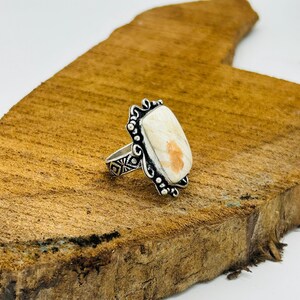 May include: A silver ring featuring an elongated rectangular stone with cream and orange marbling. The metal band displays intricate etched geometric patterns, while the stone setting is framed by decorative silver scrollwork and small beads.
