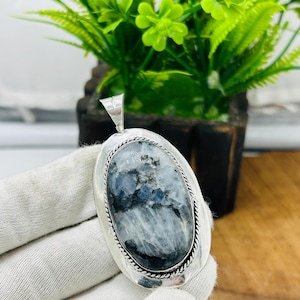 May include: An oval-shaped pendant featuring a polished moonstone with grey, white, and blue inclusions, set in a silver-toned metal frame with a twisted rope border and a triangular bail at the top for attachment to a necklace chain.