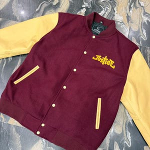 May include: A varsity jacket featuring a maroon wool-blend body and yellow sleeves. The front includes a snap-button closure, two side pockets with yellow trim, and the word "Justice" embroidered in yellow thread on the upper left chest area.