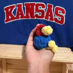 May include: A small, handmade crochet bird plushie featuring a red head, a blue body, and yellow accents on the beak and feet. The bird is positioned in front of a blue fabric background with the word KANSAS embroidered in red with white trim.